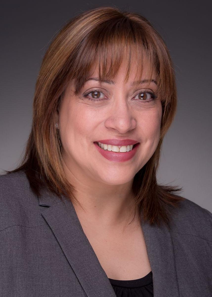 Migdaliz Berrios, a hispanic female with shoulder length brown hair is smiling and wearing a gray suit jacket with a black blouse.